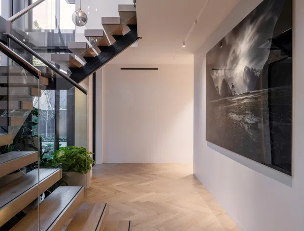 A contemporary residential stairway and hallway with floating timber stair treads, slim dark metal and glass balustrades, and a dramatic cluster of spherical pendant lights descending through the stair void. Floor-to-ceiling glazing brings in abundant daylight, while pale herringbone timber flooring continues through the open circulation space. A large monochrome artwork provides a strong feature on the white wall, with indoor planting visible beside the stairs.