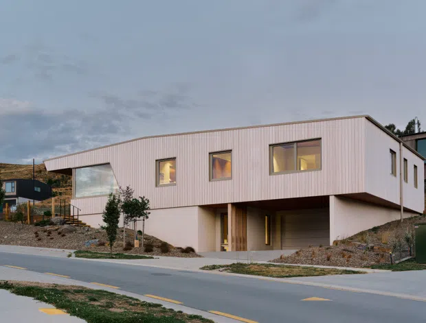 A contemporary two-storey residential house on a sloping suburban site. The upper level is clad in pale vertical ribbed panels with large rectangular windows, while the lower level has smooth light-coloured walls, a recessed carport or sheltered entry, and a timber-clad doorway. The house has a crisp cantilevered form and a restrained neutral palette, viewed from across a paved street at dusk.