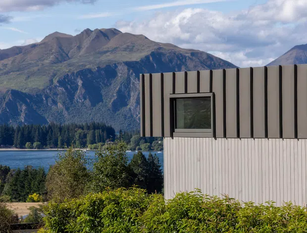 Exterior architectural view of a contemporary building beside a lake, with a vertically ribbed light facade and dark-framed projecting window. Dramatic mountains and forest form the background, while shrubs occupy the foreground.