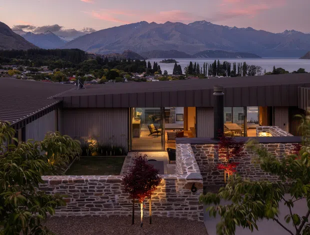 A contemporary single-storey home viewed from the landscaped exterior at dusk, with dark vertical cladding, broad glazing, a low-pitched roof and illuminated interior spaces. Dry-stone masonry garden walls and planted courtyards frame the foreground, while a lake and dramatic mountain range form the distant setting.