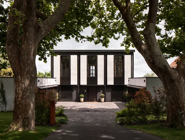 Front exterior view of a contemporary two-storey residence framed by mature trees. The house has a simple flat roof, a white and dark charcoal facade, tall narrow vertical windows and doors, and a paved driveway leading to the entrance. Low boundary walls and landscaped gardens flank the approach.