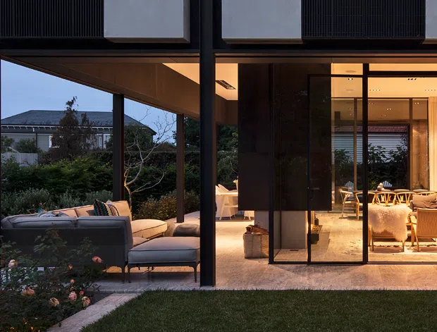 A dusk exterior view of a contemporary multi-level home, looking through a glazed living area onto a covered outdoor seating terrace. Dark structural framing, extensive floor-to-ceiling glazing, warm interior lighting, and a sheltered cantilevered upper volume define the composition. The landscaped garden and outdoor lounge are visible in the foreground, with dining and living furnishings seen through the glass.