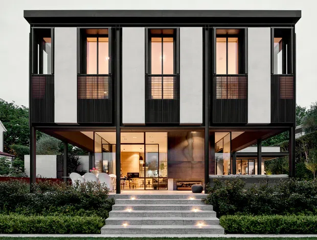 Front exterior view of a refined two-storey contemporary residence with a strongly symmetrical facade. The upper level has tall narrow windows, dark framed glazing, vertical light-coloured panels, and small screened balcony-like sections. The lower level is largely glazed and recessed beneath projecting upper volumes, revealing a warm timber-lined interior. Broad concrete steps with integrated lights lead through a manicured hedge garden to the entry.