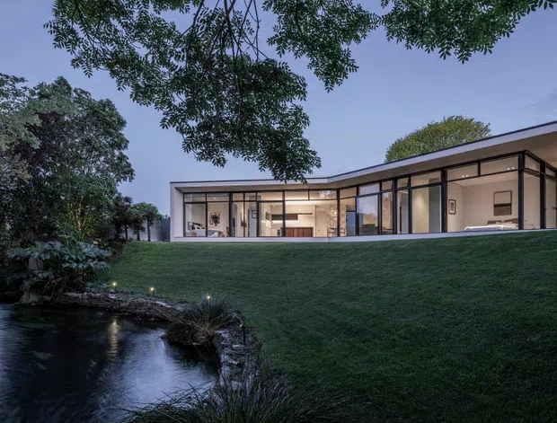 A low-slung contemporary residence at dusk, set into a landscaped sloping site beside a narrow pond or stream. The house features extensive floor-to-ceiling glazing with dark slender frames, clean white or pale rendered walls, and a shallow flat roof. Warm interior lighting reveals an open-plan living area through the windows, while mature trees, lawn, stone edging, and water create a secluded garden setting.