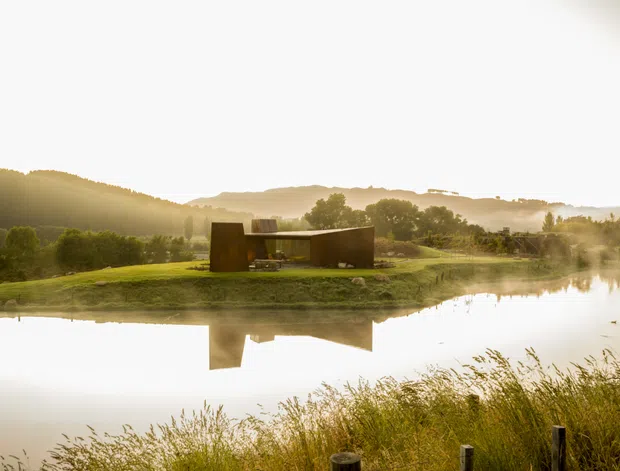 A low, minimalist architectural pavilion or house set on a grassy peninsula beside a calm reflective pond. The dark rectangular building has a strong horizontal form and appears partially embedded in the landscape, with surrounding hedges, mature trees, rolling hills, and warm low-angle sunlight creating a tranquil rural setting.