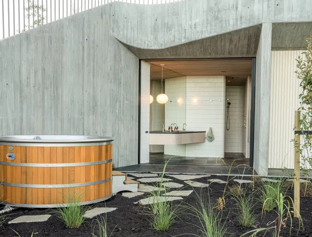 Exterior view of a contemporary concrete-clad bathroom pavilion or wet-area extension, with a curved sculptural concrete roof and wall forming a sheltered opening. Inside, a freestanding or wall-mounted curved vanity, white tiled walls, pendant globe lights, and a shower are visible. A round timber-clad outdoor spa sits in the landscaped foreground, surrounded by dark gravel, grasses, and stepping stones.