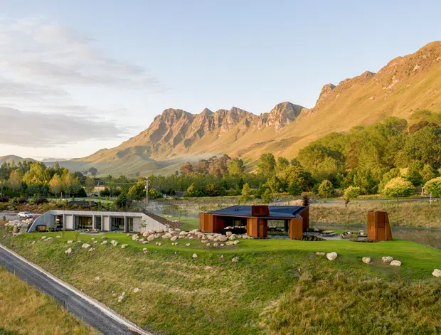 A wide exterior view of a low-profile contemporary residential property set in a landscaped rural valley. The buildings use dark timber and neutral cladding, with extensive glazing and a flat or very low-pitched roofline. A paved driveway, lawn, rock landscaping, trees, and a narrow waterway sit in the foreground, while steep rugged mountains dominate the background.