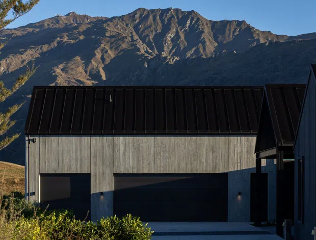 Exterior view of a contemporary dark-clad building with a steep metal roof and vertically textured concrete or grey cladding, set against a dramatic rugged mountain backdrop. The foreground includes landscaped shrubs and a driveway or paved approach, while adjoining dark vertical siding is visible on the right.