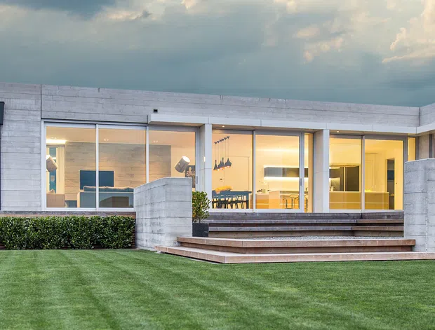 Wide exterior view of a low-slung contemporary residence with a flat roof, pale concrete masonry walls, extensive floor-to-ceiling glazing, and broad timber steps opening onto a manicured lawn. Warm interior lighting is visible through the windows, with distant hills and dramatic cloudy sky beyond the property.