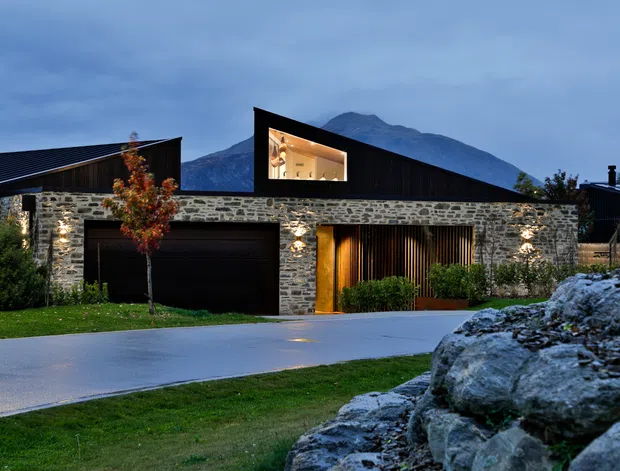 Contemporary single-storey residence with a dark pitched roof, low stone-clad facade, integrated garage, and warm timber-screened entry. Large upper clerestory glazing reveals a warmly lit interior, while landscaped lawn and rocks frame the home against a mountain backdrop at dusk.