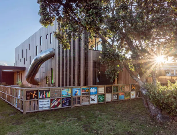 Exterior view of a contemporary multi-level building with vertical timber-look cladding, narrow slit windows, and a large curved metal slide projecting from an upper level. A colourful artwork-decorated timber fence encloses a grassy outdoor area, with mature trees and strong afternoon sunlight surrounding the building.