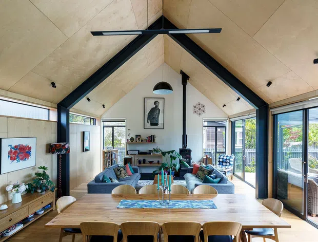A bright open-plan living and dining room beneath a dramatic pitched, timber-lined ceiling with exposed dark structural beams. The room features a large timber dining table, upholstered dining chairs, a blue sectional seating area, a central fireplace or flue, extensive glazing and sliding doors opening to an outdoor terrace. The interior combines natural timber, black framing, contemporary furnishings and abundant daylight.
