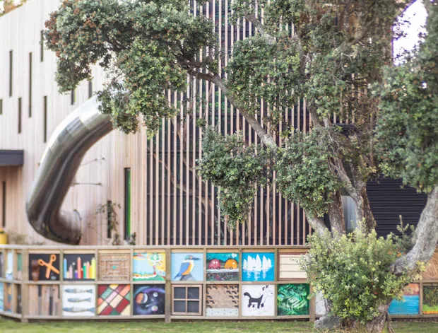 Exterior view of a contemporary building with pale vertical cladding, a tall dark vertical-slat facade screen, and a prominent polished metal tube-like architectural feature. Mature trees partially screen the facade, while a low timber-framed wall displays colourful artwork or panels along the foreground.