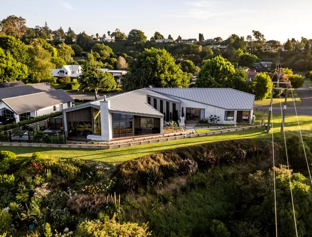 Aerial view of a contemporary low-rise residential property in a lush, landscaped suburban or semi-rural setting. The complex includes several light-coloured buildings with pitched metal roofs, extensive glazing, covered outdoor areas, timber decking, lawns, and established gardens. A road and neighbouring homes are visible in the background.