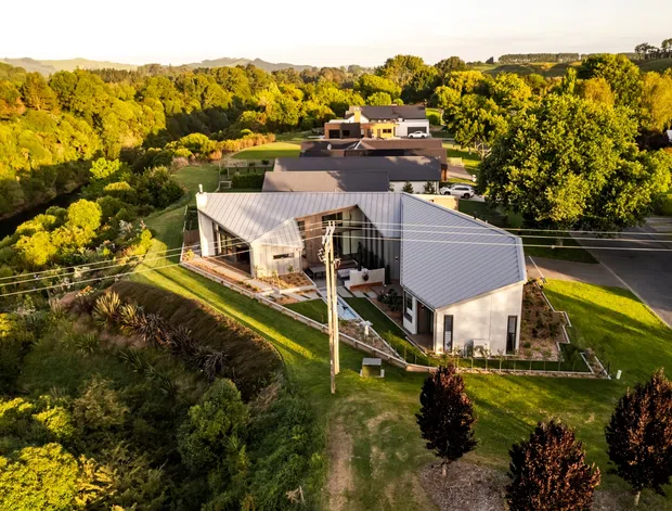 Aerial view of a contemporary low-rise residential property in a lush, tree-filled suburban or semi-rural setting. The main building has multiple connected wings, light grey metal-clad pitched roofs, pale exterior walls, large glazed openings, and landscaped lawns. Roads, neighbouring homes, overhead utility lines, and extensive greenery are visible around the site.
