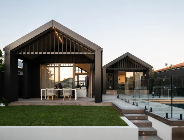 Exterior view of a contemporary single-storey residence with two dark gabled pavilion forms, dramatic vertical timber-style facade screening, extensive glazing, and a covered outdoor dining area. A glass pool fence, swimming pool, lawn, paving, and stepped terrace connect the building to the landscaped outdoor space.