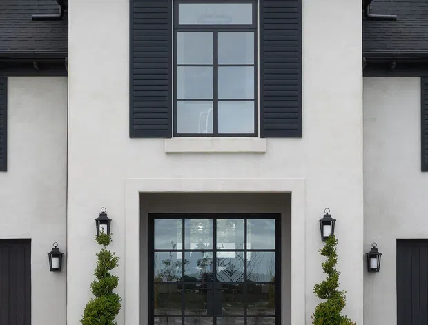 Symmetrical front facade of a refined two-storey residence, featuring pale rendered walls, a dark pitched shingle roof, black-framed multi-pane glazing, matching louvered shutters, traditional lantern sconces, and formal topiary planters flanking the recessed entry.