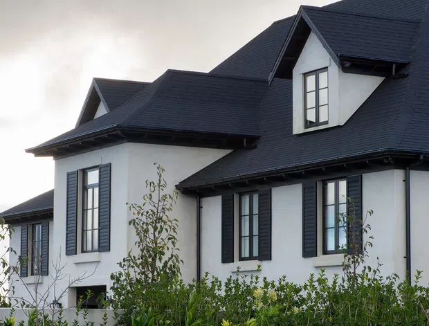 Exterior view of a large multi-gabled house with dark pitched shingle roofing, white rendered walls, black-framed windows, and dark louvered shutters. Landscaping partially screens the lower facade.