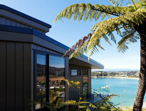 Exterior view of a dark-clad, low-profile modern residence on a steep, vegetated site overlooking a bright blue coastal inlet or bay. The building has angular rooflines, large dark-framed windows, and a deck or stair structure descending toward the water. Ferns and a tall tree dominate the foreground, with a settled shoreline and hills visible across the inlet.