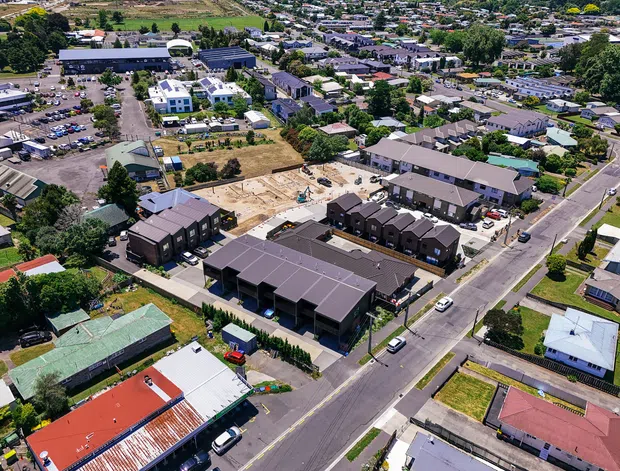 Aerial view of a suburban mixed residential and commercial area, with recently constructed dark-roofed townhouses and apartment-style buildings surrounding an active construction site. Low-rise housing, streets, parking areas, community facilities, and landscaped sections are visible across the neighbourhood.
