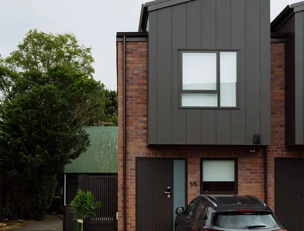 Street-facing exterior view of a contemporary attached townhouse. The facade combines red brick side walls with dark charcoal vertical metal cladding, projecting upper-level volumes, modern rectangular windows, and a dark garage and entry door. A concrete driveway, landscaped narrow garden bed, neighbouring homes, and a parked car are visible.