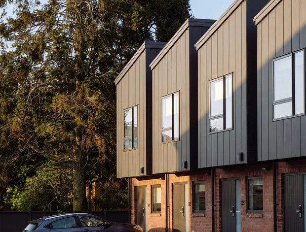 Street-level exterior view of a row of contemporary attached townhouses. The buildings feature steeply angled gabled roof forms, dark vertical metal cladding, warm brown vertical-panel cladding, exposed red-brick lower façades, tall rectangular windows, and integrated garages. A landscaped driveway with small shrubs and a parked vehicle is visible in front, with mature trees behind the development.