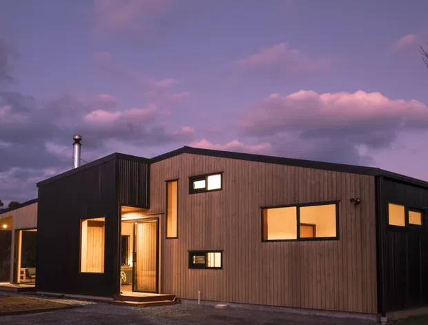 A modern single-storey residence photographed at dusk, featuring a simple flat-roofed and low-pitched roof composition, dark vertically ribbed cladding, warm illuminated windows, and a sheltered entry porch. The home sits in a landscaped, tree-lined setting with a dramatic purple evening sky.
