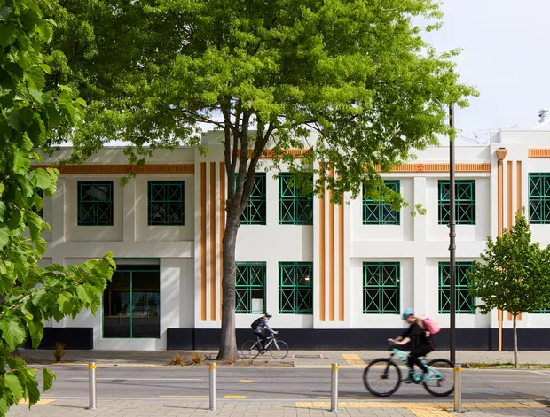 Street-facing view of a two-storey institutional or commercial building with a white rendered facade, strong orange vertical accents and cornice detailing, and repeated dark green geometric window grilles. Mature street trees and cyclists occupy the foreground, with bollards and a paved streetscape along the building.