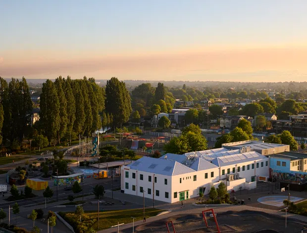 Elevated wide view of a suburban urban area at sunset, with a prominent cluster of low-rise contemporary buildings in the foreground, surrounding roads, a playground, tall trees, and residential development extending into the distance.