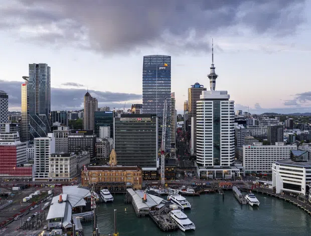 Aerial cityscape of a dense waterfront central business district, with high-rise office and apartment towers, a prominent communications tower, and a busy harbour ferry terminal with multiple piers and vessels. The image is photographed from above and focuses on the urban waterfront skyline and built environment under a cloudy sky.