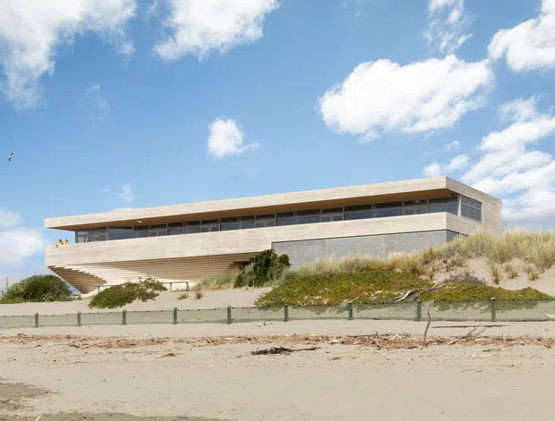 A contemporary beachfront building viewed from the sandy shore, composed of long horizontal volumes with pale timber-toned cladding, extensive narrow clerestory glazing, and broad projecting roof slabs. Dunes and coastal grasses surround the structure beneath a bright blue sky.