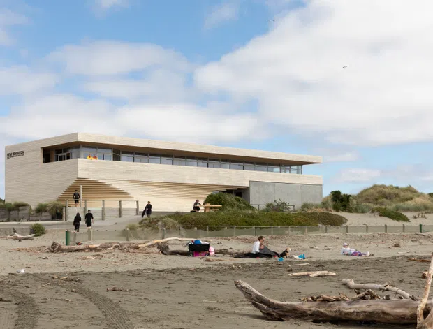 A contemporary two-storey coastal building viewed from a sandy beach. The structure has a long, low horizontal form, pale horizontal cladding, extensive upper-level glazing, a projecting flat roof, and a prominent exterior stair. Dune vegetation, driftwood, beachgoers, and a wide sky surround the building.