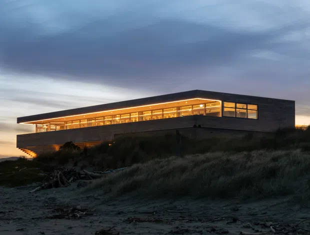 A modern, low-profile coastal building viewed at dusk from a sandy, grass-covered site. The dark masonry or cladding exterior has a long projecting flat roof, extensive warm-lit glazing, and a dramatic cantilevered upper volume integrated into the dune landscape.