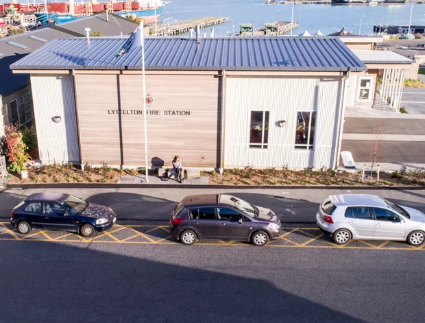 Exterior aerial view of the Lyttelton fire station, a compact modern single-storey building with a ribbed metal gable roof, pale vertical cladding, and white-framed windows and doors. It fronts a paved street with parked cars and overlooks a marina and industrial waterfront.