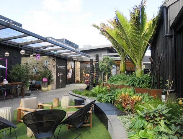A lush landscaped courtyard at a contemporary commercial venue, with outdoor café seating, artificial turf, raised garden beds, tropical palms and dense planting. Dark vertical cladding, exposed steel framing and translucent roof panels create a sheltered outdoor dining and lounge environment, with exposed brick and signage along the surrounding walls.