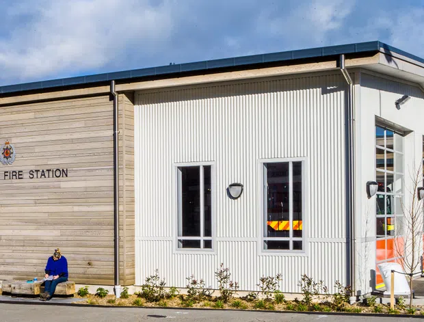 Exterior view of the Lyttelton Fire Station, showing a contemporary civic building with horizontal timber-look cladding, vertically ribbed light facade panels, tall windows, a low-pitched roof, and visible fire-station equipment through the glazing. A flagpole, signage, entrance, and landscaped frontage are also visible.