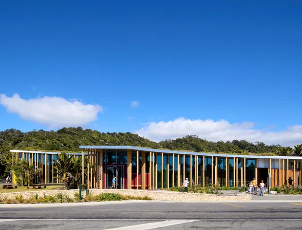Low-rise contemporary building viewed from across a road, featuring long horizontal glazed facades, slender vertical timber or metal fins, warm timber-clad sections, and broad flat roof planes. The building sits within landscaped grounds against a wooded backdrop under a clear blue sky.
