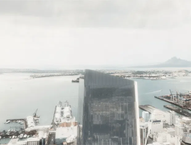 Wide aerial view of a dense waterfront city district, dominated by a tall dark glass high-rise in the foreground. A broad harbour, piers, boats, and distant low-rise urban development extend across the background beneath an overcast sky.