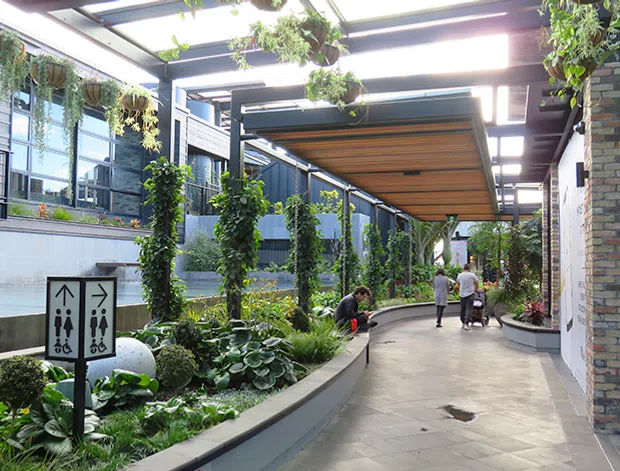 A covered landscaped pedestrian courtyard or walkway in a contemporary building complex, with extensive hanging and climbing greenery, planted garden beds, concrete paving, dark metal and glass structures, a timber-lined canopy, and exposed multicoloured brick walls. Directional restroom signage and people are visible in the distance.