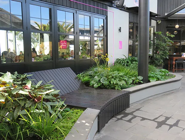 A contemporary indoor-outdoor commercial courtyard or atrium with dark metal-framed glazing, exposed structural columns and roof elements, lush integrated planting, built-in curved timber bench seating, and a paved circulation area. A café or hospitality seating area is visible in the background through the open space.