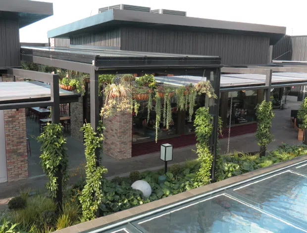 Exterior view of a contemporary commercial venue beside a swimming pool, featuring dark vertical cladding, flat roof forms, covered outdoor dining or walkway structures, hanging greenery, planted landscaping, and brick masonry walls. The pool occupies the foreground.