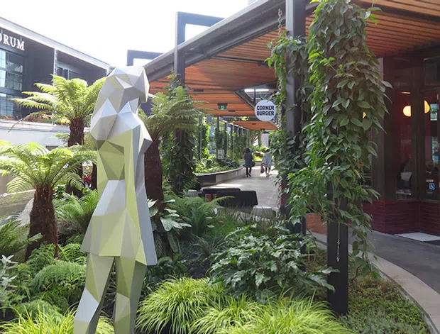 A landscaped pedestrian courtyard or outdoor dining precinct with lush tropical planting, a faceted geometric human sculpture, timber-lined covered structures, and glazed storefronts. The image includes a visible restaurant or commercial venue entrance and signage, with modern angular architecture and shaded walkways.