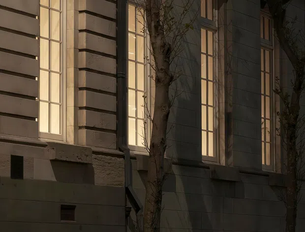 A close-up exterior view of a refined, classical-style building facade at dusk. Tall narrow multi-pane windows glow warmly within a pale stone or rendered wall, framed by projecting pilasters, horizontal banding, cornices and deep window surrounds. Slender trees and low planted greenery line the base, while dramatic side lighting creates a moody, elegant atmosphere.