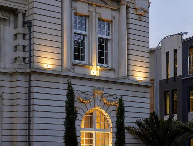 Street-level exterior view of a grand neoclassical-style building with a pale grey stone or rendered facade, strong horizontal rustication, ornate classical columns and capitals, decorative cornice, arched window surround, and tall multi-pane windows glowing with warm interior light. A contemporary neighbouring building, planted frontage, and parked cars are also visible.