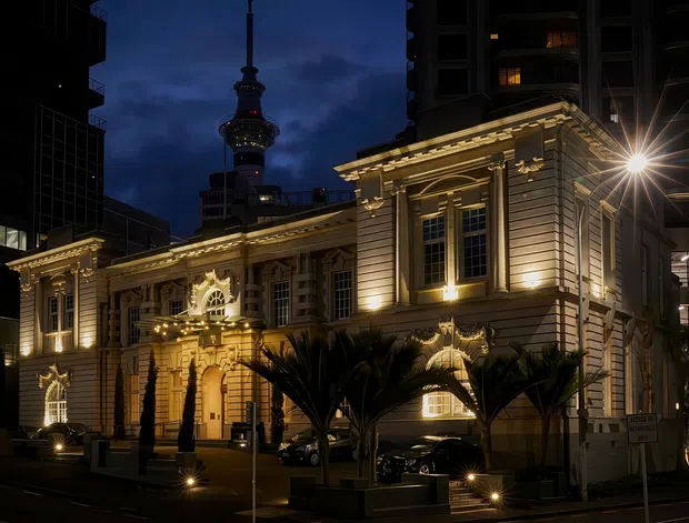 Night-time exterior photograph of an ornate historic civic or commercial building illuminated with warm architectural lighting. The symmetrical facade features classical detailing, pilasters, cornices, tall multi-pane windows, decorative reliefs, and a grand central entrance. Modern high-rise buildings and a distinctive illuminated tower rise behind it, while palm-like plantings line the street frontage.