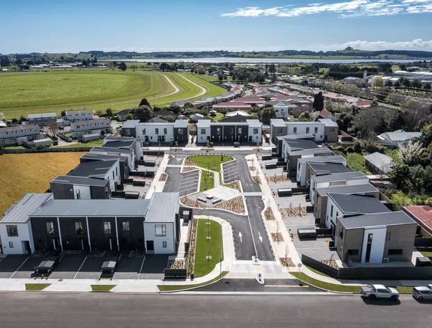 Aerial view of a newly developed residential neighbourhood with multiple modern, predominantly single-storey homes arranged along landscaped streets and a central green space. The development is bordered by open agricultural fields, waterways and established residential areas, with a clear suburban master-planned layout.