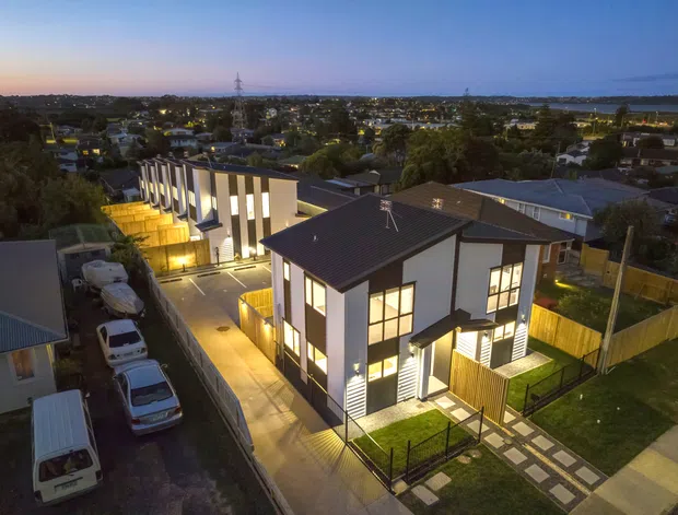 Aerial dusk view of a contemporary multi-unit residential development, with several compact two-storey homes featuring dark pitched roofs, white rendered walls, vertical cladding accents, timber fencing, landscaped paths and shared driveways. A suburban neighbourhood and distant waterfront or coastline are visible beyond the development.