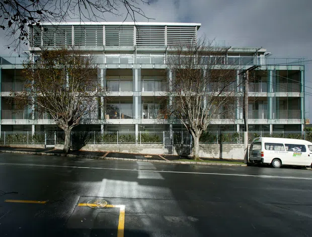 Street-facing view of a contemporary multi-storey residential or mixed-use building with a rectilinear glass-and-concrete facade, balconies, extensive horizontal louvres, and a flat roof. Mature street trees and parked vehicles sit in front of the building along a wet urban roadway.