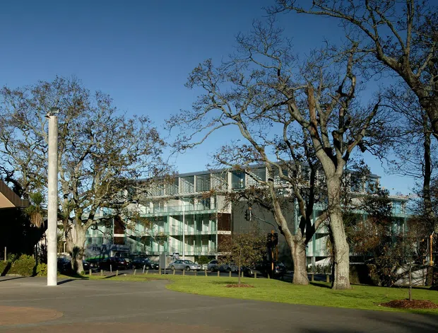 Exterior architectural view of a contemporary institutional or commercial complex. A broad low-slung building with a sharply projecting roof canopy and slender white columns occupies the left, while a multi-storey glazed building is visible beyond. Mature deciduous trees, open paved forecourt, lawn, and clear blue sky define the setting.