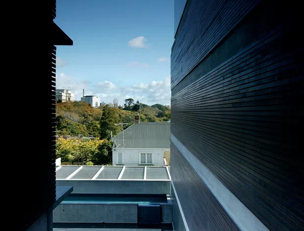 Vertical architectural exterior view looking through a narrow urban courtyard or gap between buildings. The dominant facade on the right is clad in dark horizontal timber boards, with contrasting pale concrete or rendered bands and reflective glazing. A lower-level roof terrace or courtyard with a dark rectangular water-like surface is visible below, while neighbouring residential buildings and landscaped vegetation appear in the distance. The image has strong vertical perspective, deep shadows, and cool blue daylight.